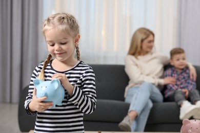 Photo of Family budget. Little girl putting coin into piggy bank while her mother and brother sitting on sofa at home, selective focus