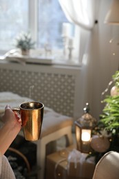 Photo of Woman with golden cup of hot drink in room decorated for Christmas, closeup