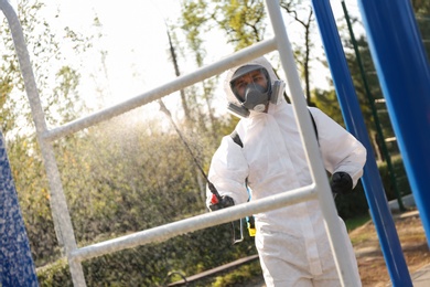 Photo of Man in hazmat suit spraying disinfectant on outdoor gym's equipment. Surface treatment during coronavirus pandemic