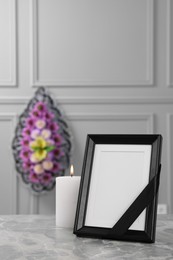 Photo of Photo frame with black ribbon, burning candle on light grey table and wreath of plastic flowers near wall indoors. Funeral attributes