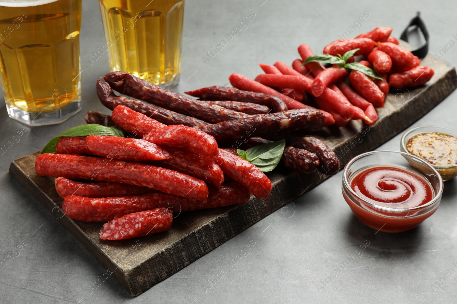 Photo of Different thin dry smoked sausages, sauces and glasses of beer on grey table