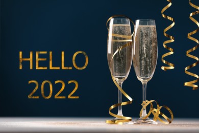 Image of Hello 2022. Glasses of sparkling wine and serpentine streamers on table against dark blue background 