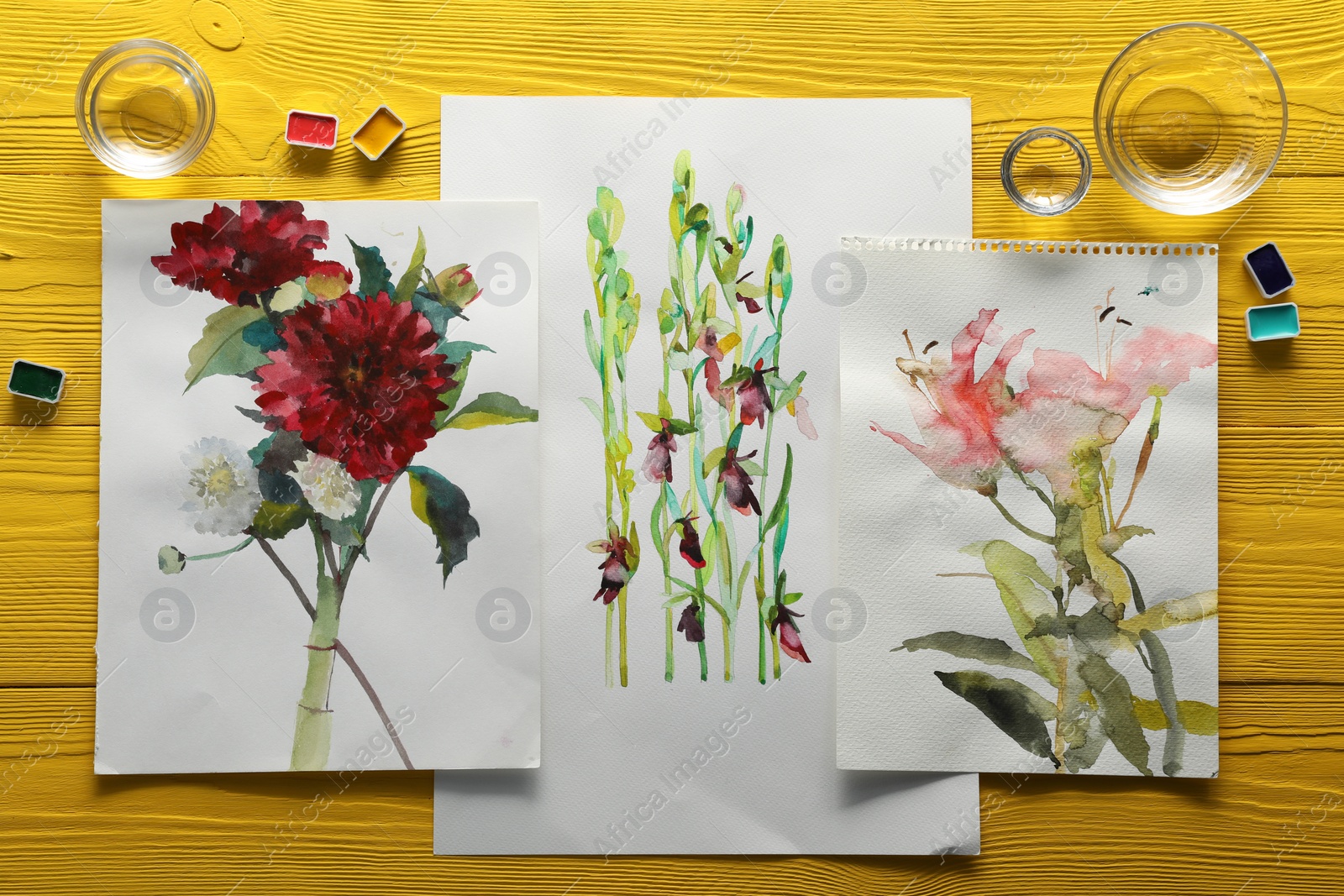 Photo of Beautiful floral pictures and watercolor paints on yellow wooden table, flat lay