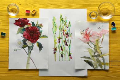 Beautiful floral pictures and watercolor paints on yellow wooden table, flat lay