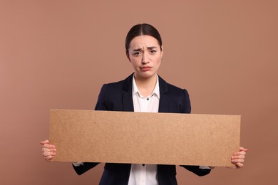 Photo of Upset young woman holding blank cardboard banner on brown background, space for text