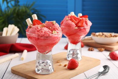 Photo of Delicious scoops of strawberry ice cream with wafer sticks and nuts in glass dessert bowls served on white wooden table, closeup