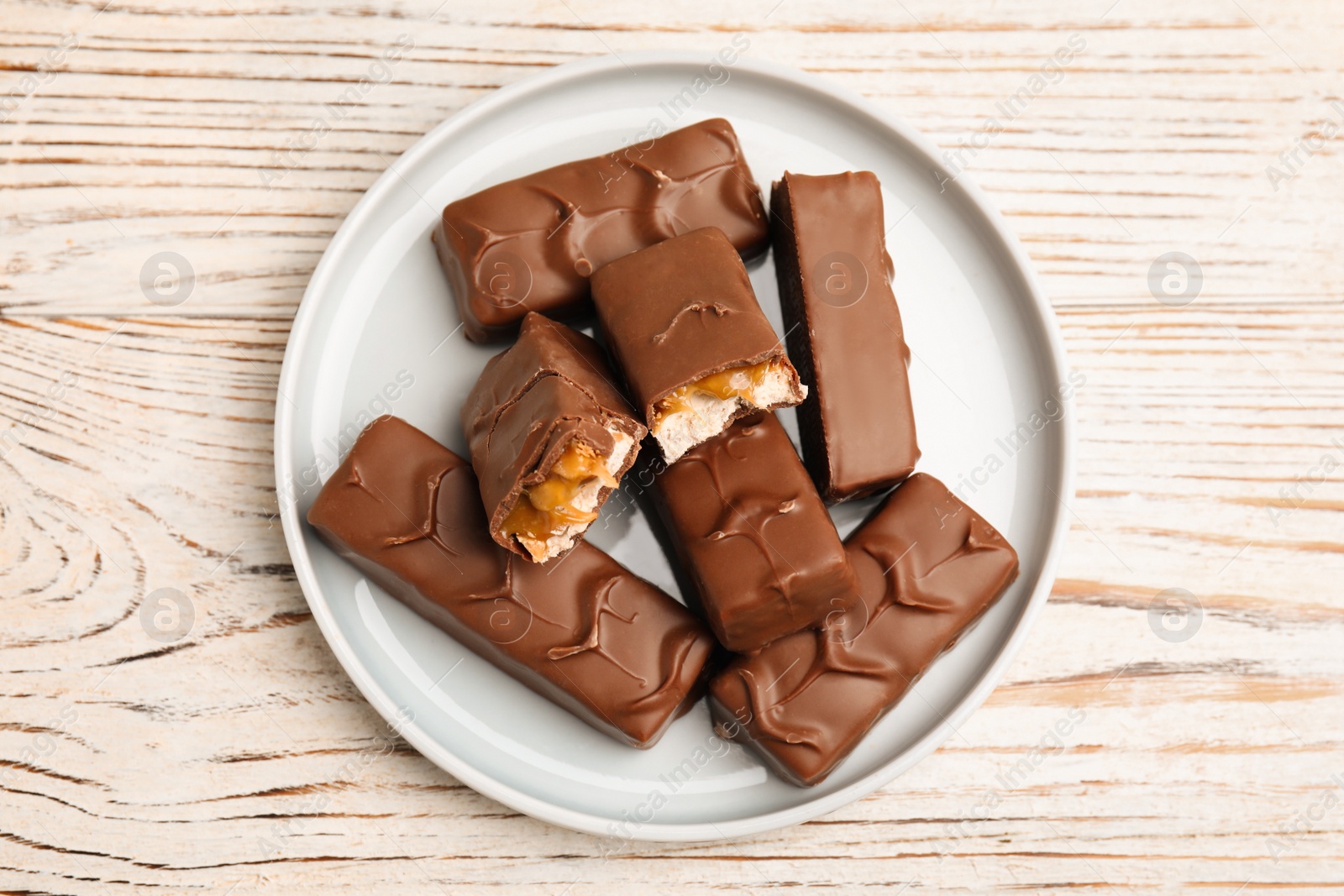 Photo of Plate of chocolate bars with caramel, nuts and nougat on white wooden table, top view