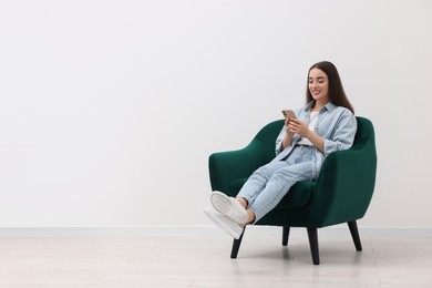 Photo of Beautiful woman using smartphone while sitting in armchair near white wall indoors, space for text