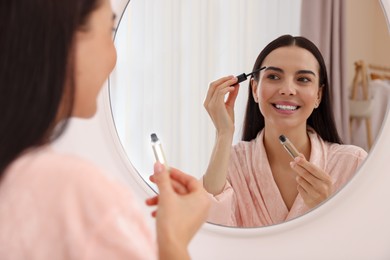 Beautiful young woman applying eyebrow gel near mirror indoors