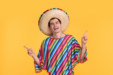 Young man in Mexican sombrero hat and poncho pointing at something on yellow background