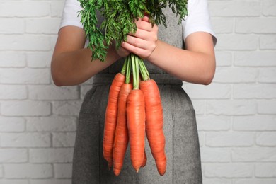 Photo of Woman holding fresh ripe juicy carrots against white brick wall, closeup