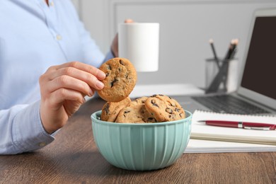 Office worker with cup of drink taking chocolate chip cookie from bowl at workplace, closeup