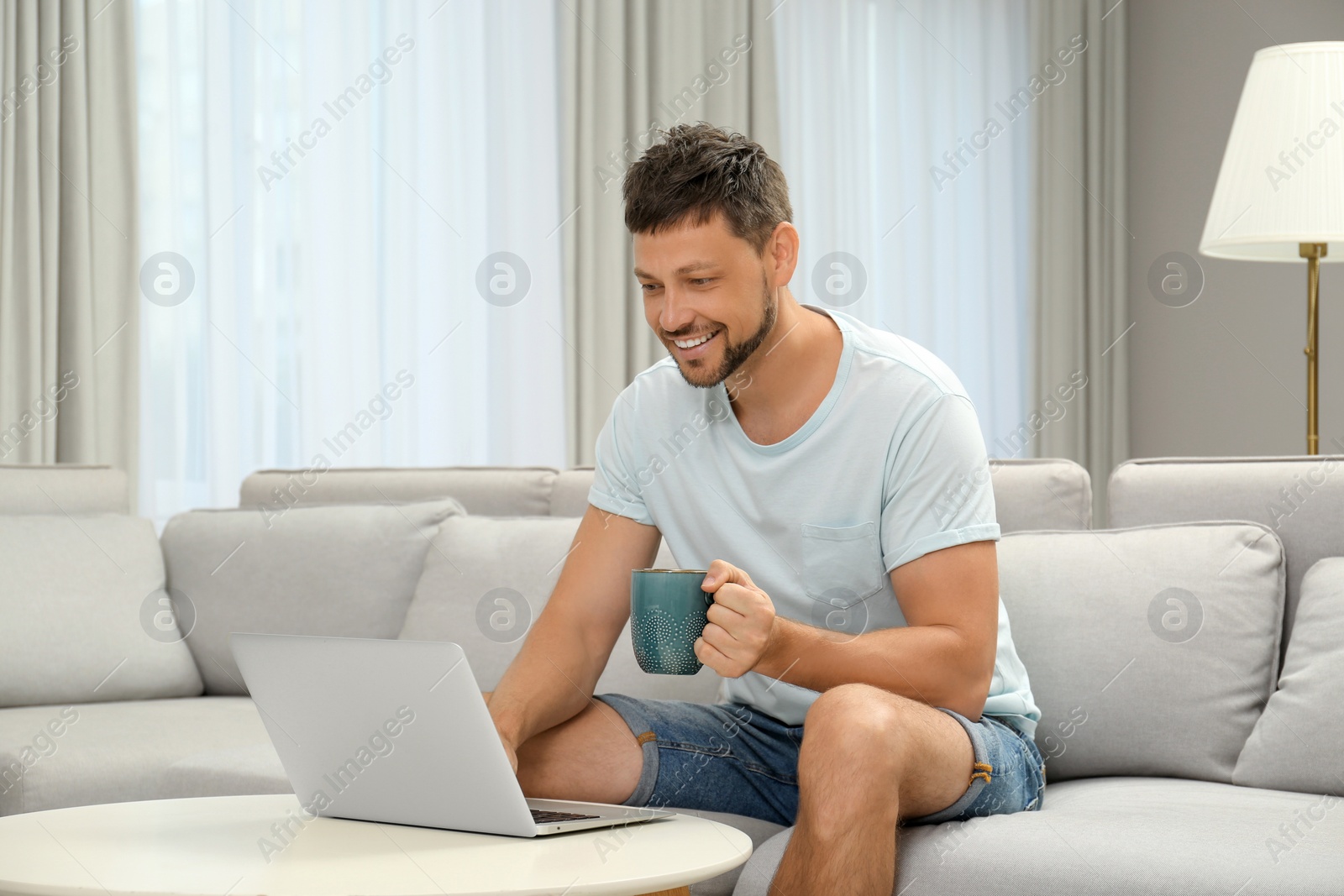 Photo of Happy man with cup of drink using laptop on sofa at home. Internet shopping