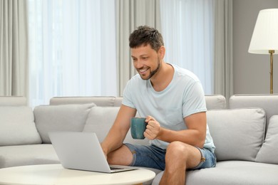 Happy man with cup of drink using laptop on sofa at home. Internet shopping