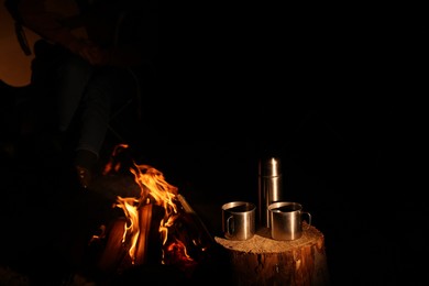 Photo of Man with guitar near bonfire at night, focus on metal mugs and thermos
