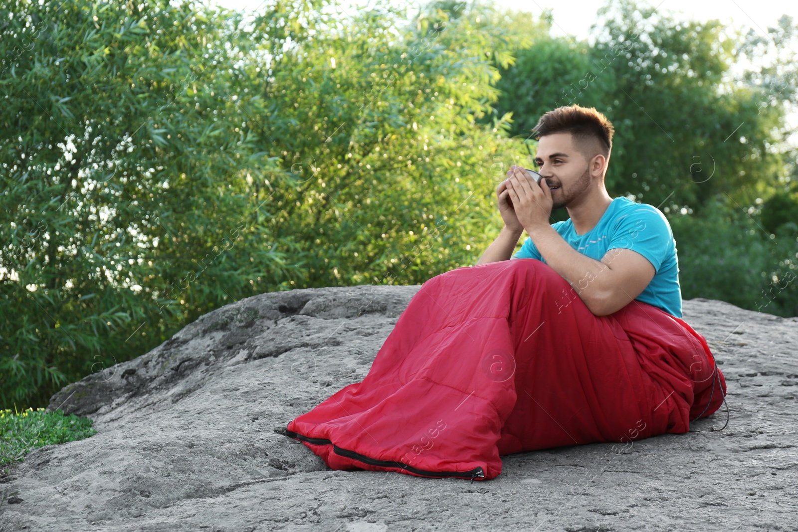 Photo of Young man with cup in sleeping bag sitting on rock. Space for text