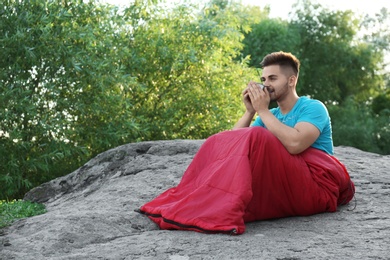 Young man with cup in sleeping bag sitting on rock. Space for text