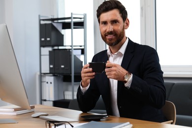 Photo of Smiling bearded man with cup of drink at table in office