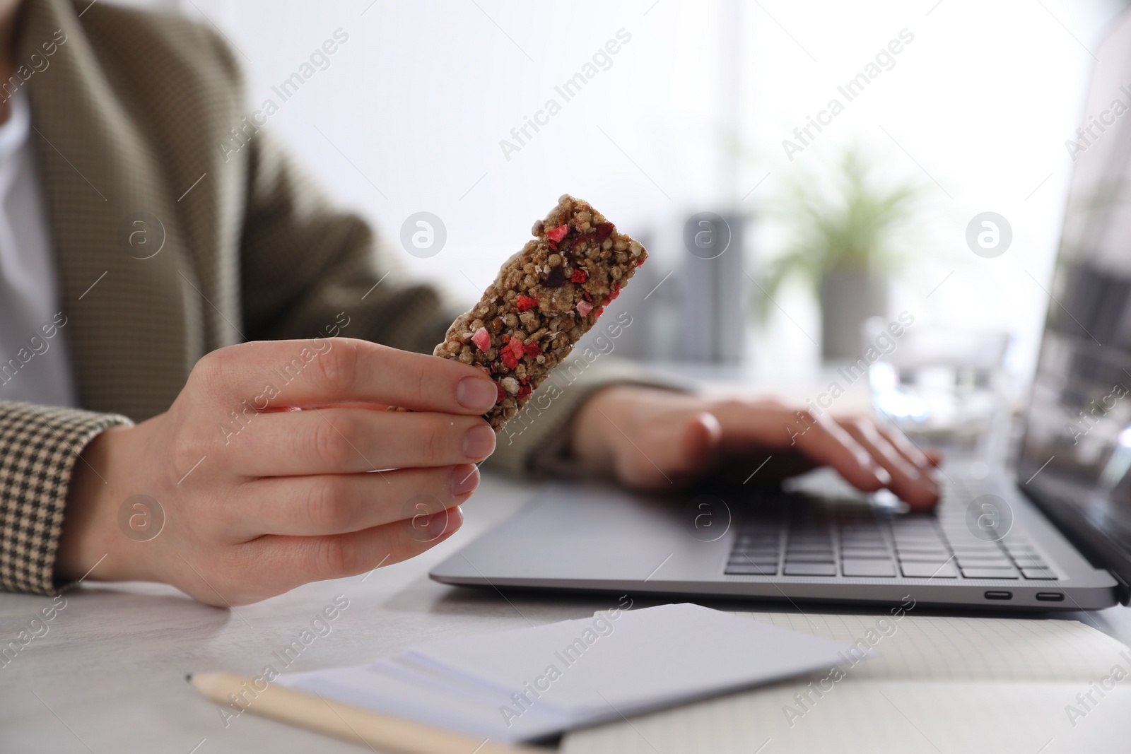 Photo of Woman holding tasty granola bar working with laptop at light table in office, closeup