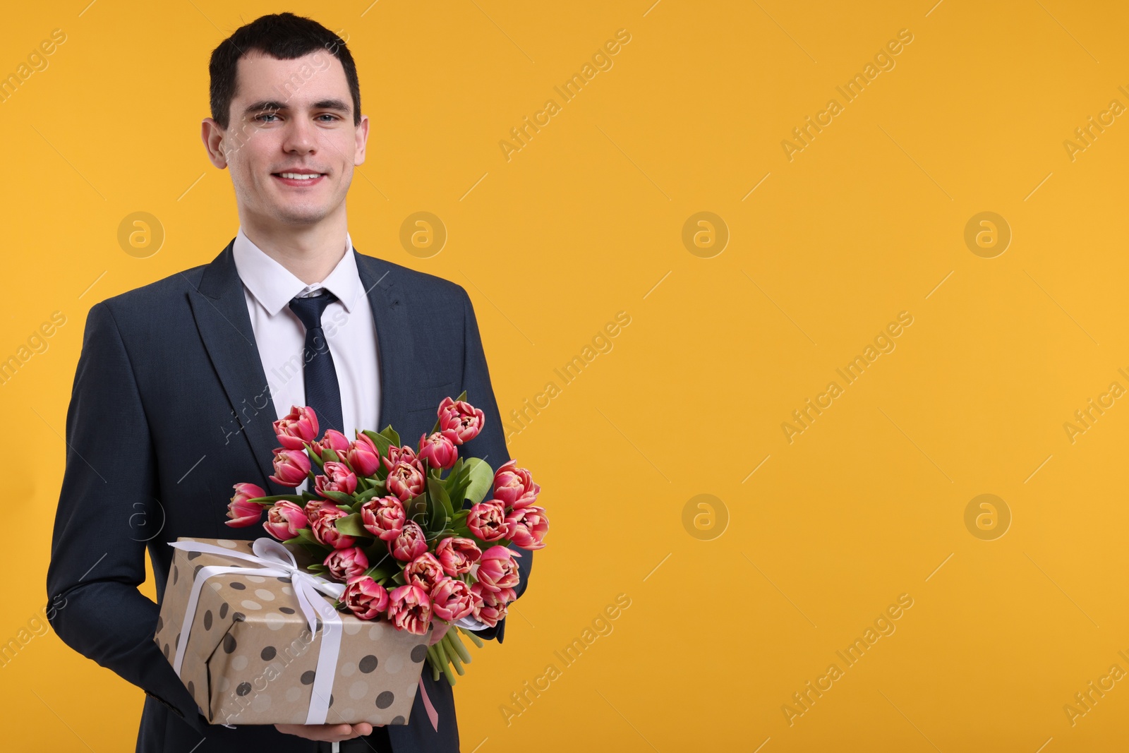 Photo of Happy young man with beautiful bouquet and present on orange background. Space for text