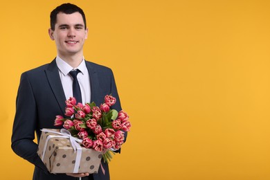 Photo of Happy young man with beautiful bouquet and present on orange background. Space for text