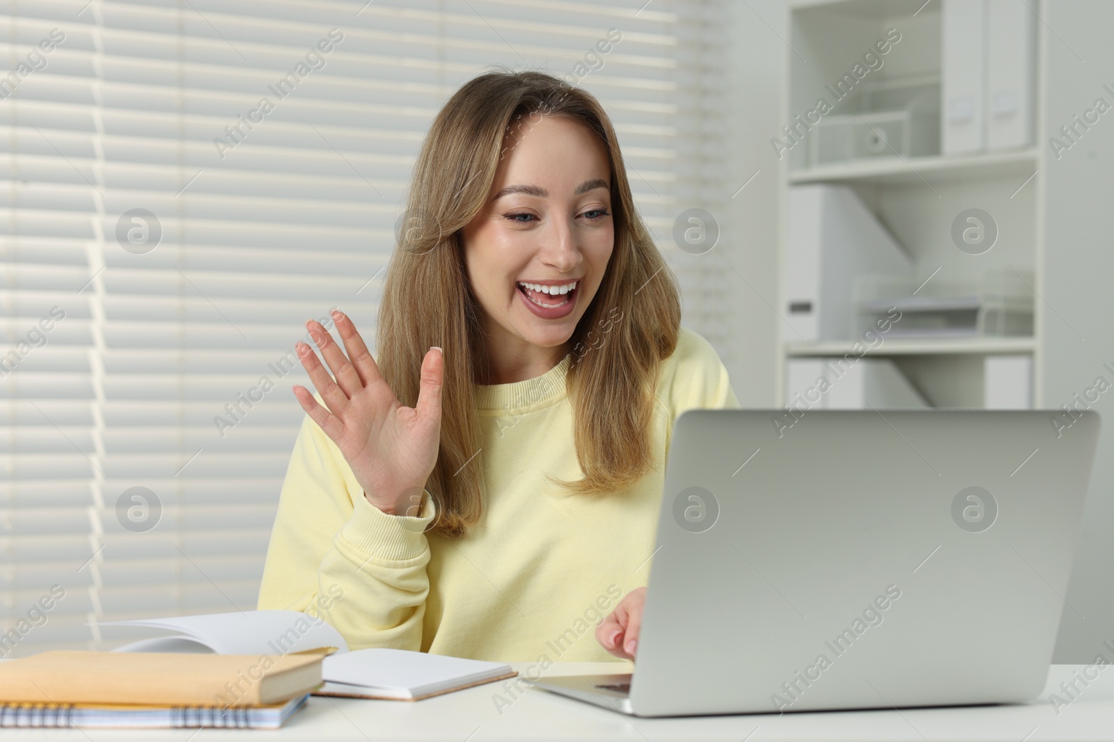 Photo of Young woman waving hello during video chat via laptop at white table indoors