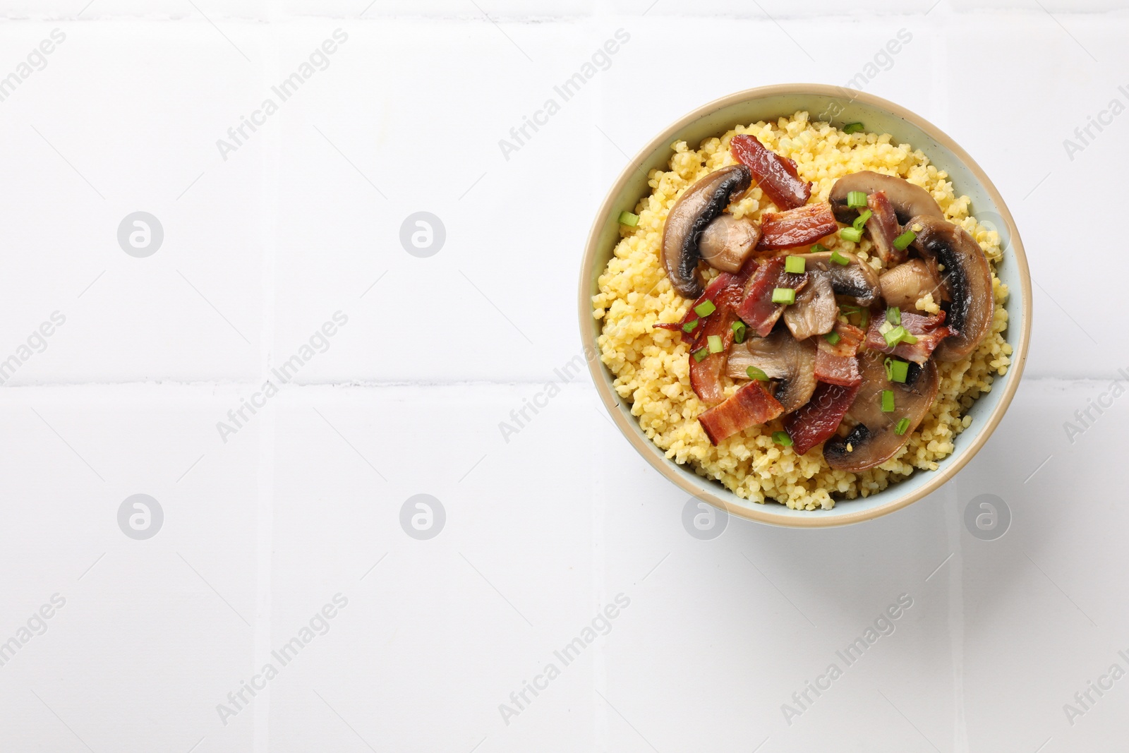 Photo of Tasty millet porridge with mushrooms, bacon and green onion in bowl on white tiled table, top view. Space for text