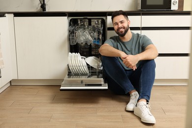 Photo of Smiling man sitting near open dishwasher in kitchen