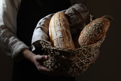 Photo of Man holding wicker basket with different types of bread on dark brown background, closeup