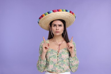 Young woman in Mexican sombrero hat pointing at something on violet background