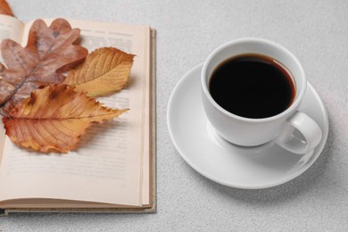Cup of hot drink, book and autumn leaves on light grey textured table