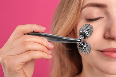 Young woman using metal face roller on pink background, closeup