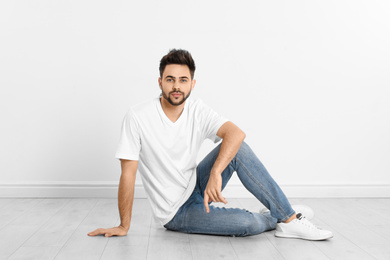 Young man in stylish jeans sitting near white wall