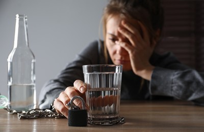 Photo of Alcohol addiction. Woman chained with glass of vodka at wooden table in room, focus on hand