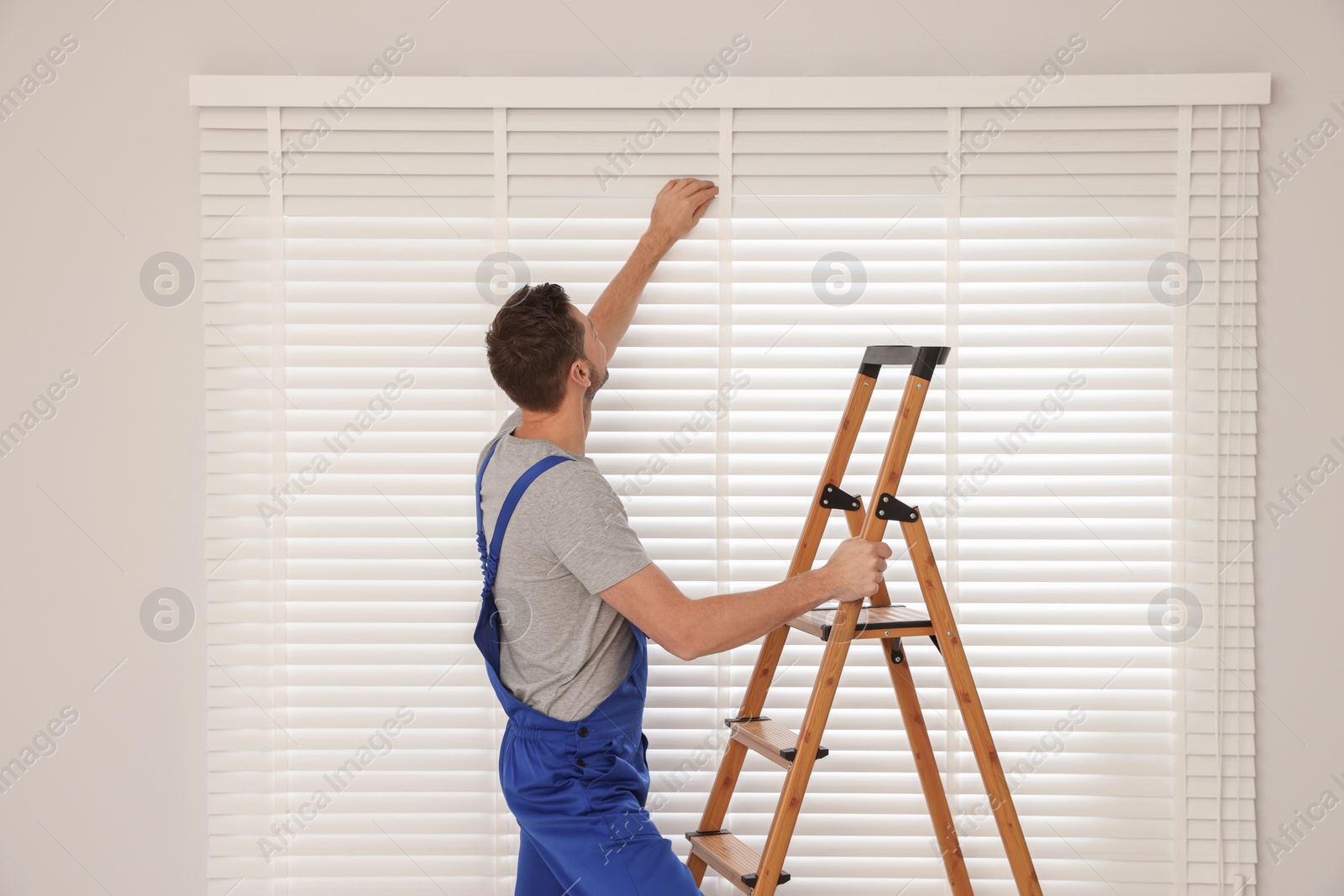 Photo of Worker in uniform installing horizontal window blinds on stepladder indoors