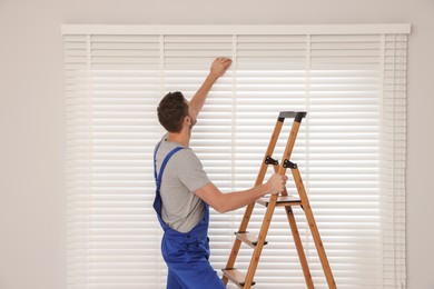 Worker in uniform installing horizontal window blinds on stepladder indoors