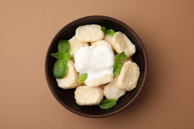 Bowl of tasty lazy dumplings with sour cream and mint leaves on light brown background, top view