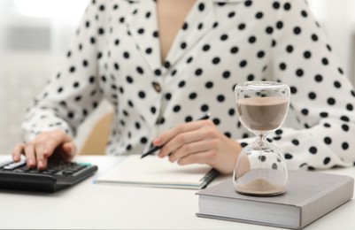 Photo of Hourglass with flowing sand on desk. Woman taking notes while using calculator indoors, selective focus