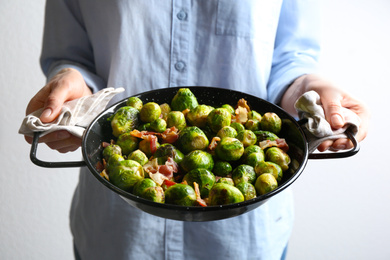 Photo of Woman holding wok with Brussels and bacon on light background, closeup