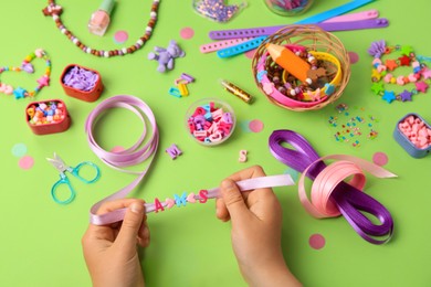 Child making beaded jewelry and different supplies on green background, above view. Handmade accessories