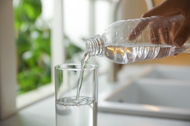 Woman pouring water from bottle into glass in kitchen, closeup