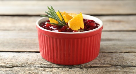 Fresh cranberry sauce in bowl, rosemary and orange peel on wooden table, closeup