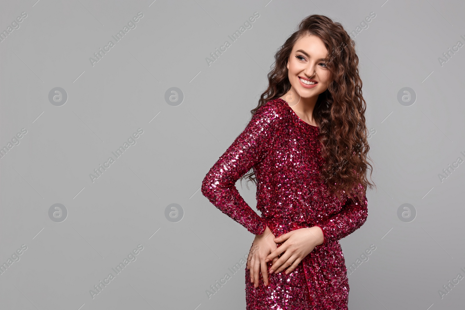 Photo of Beautiful young woman with long curly brown hair in pink sequin dress on grey background, space for text