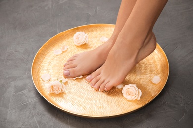 Photo of Woman soaking her feet in plate with water and roses on grey background, closeup. Spa treatment