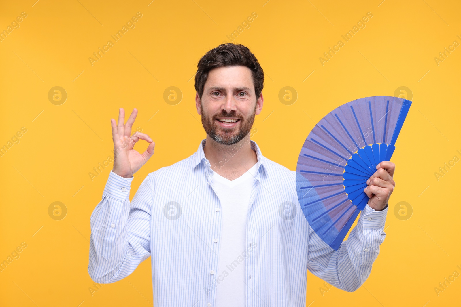 Photo of Happy man holding hand fan and showing ok gesture on orange background