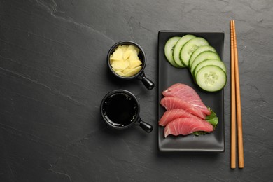 Photo of Tasty sashimi (pieces of fresh raw tuna), sliced cucumber, soy sauce and ginger slices on black table, flat lay. Space for text