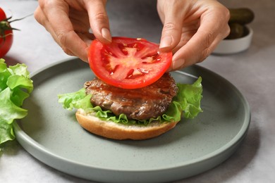 Photo of Woman making tasty hamburger with fried patty, vegetables and bun at table, closeup