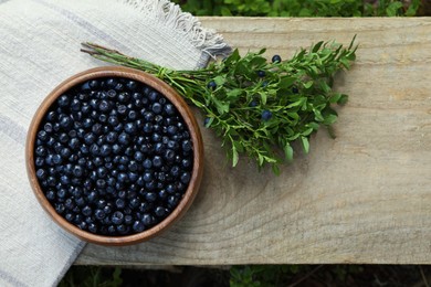 Delicious bilberries in bowl and branch with fresh berries on wooden bench, flat lay