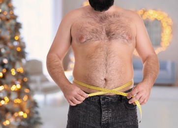 Overweight man measuring his waist in room with Christmas tree after holidays, closeup