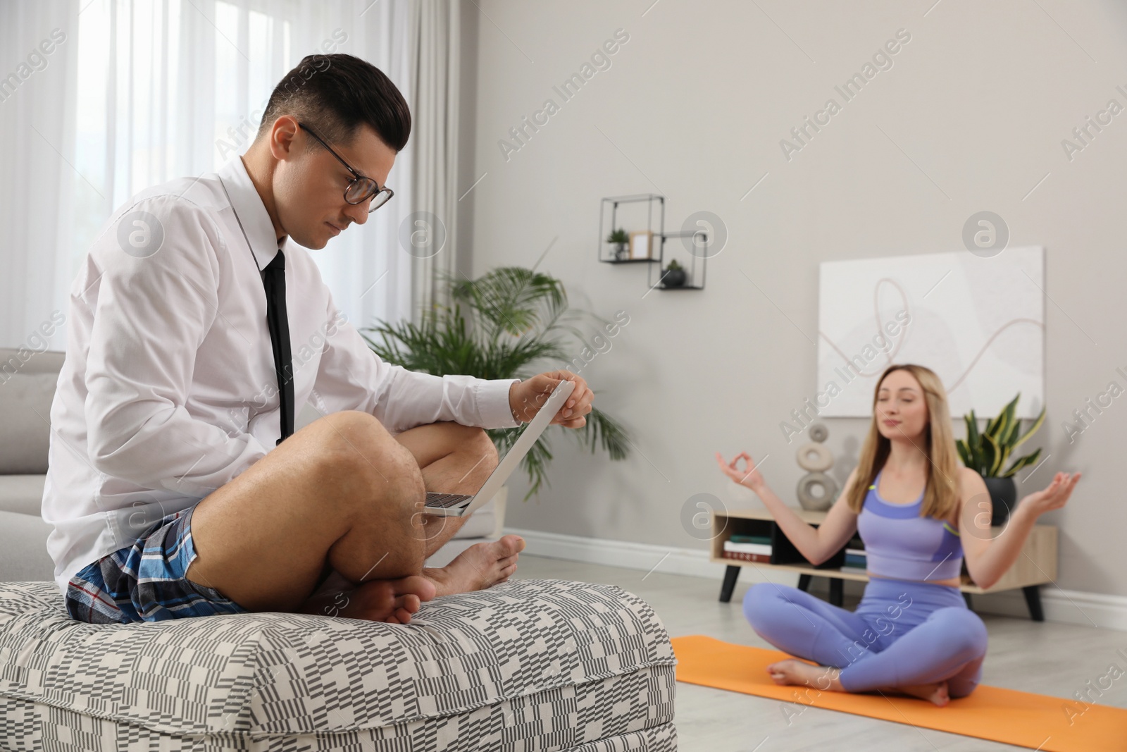 Photo of Man working on laptop while his wife practicing yoga in living room. Stay at home concept